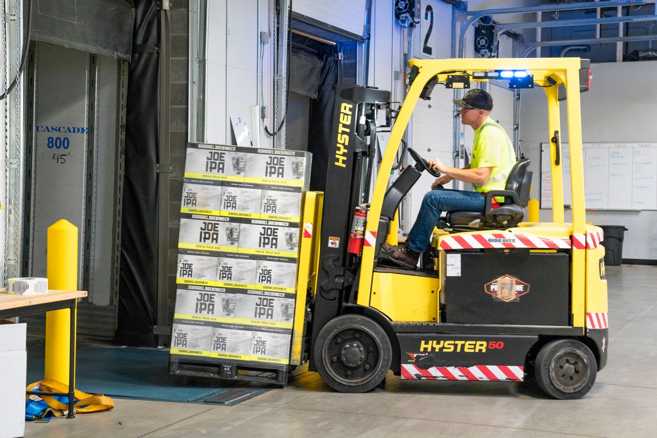 A worker drives a Hyster forklift moving Joe IPA boxes in a warehouse.